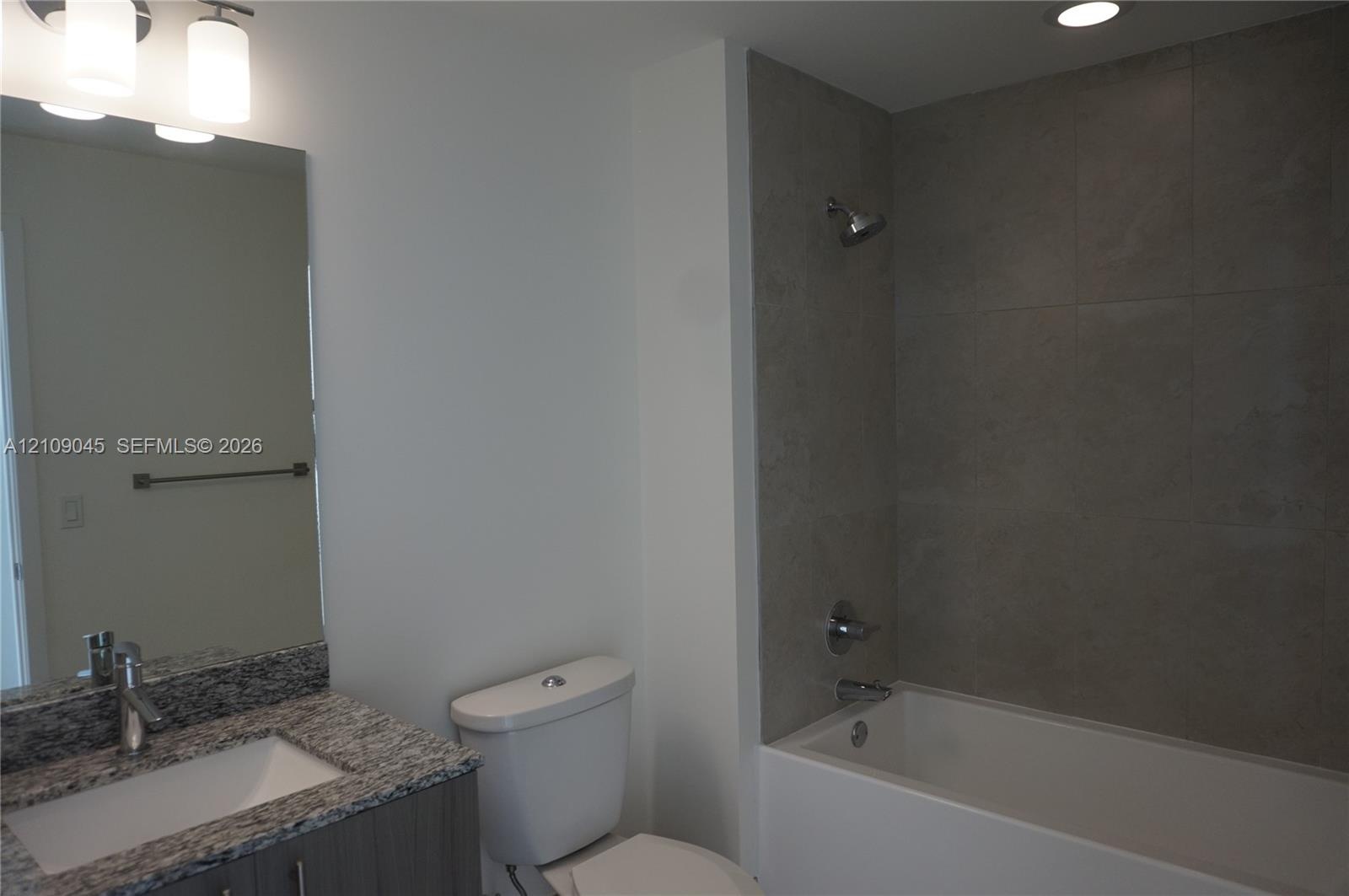 Bathroom featuring a vanity with a stone-finish countertop and integrated sink, chrome faucet, large frameless mirror, and a tub with a tile-surround 