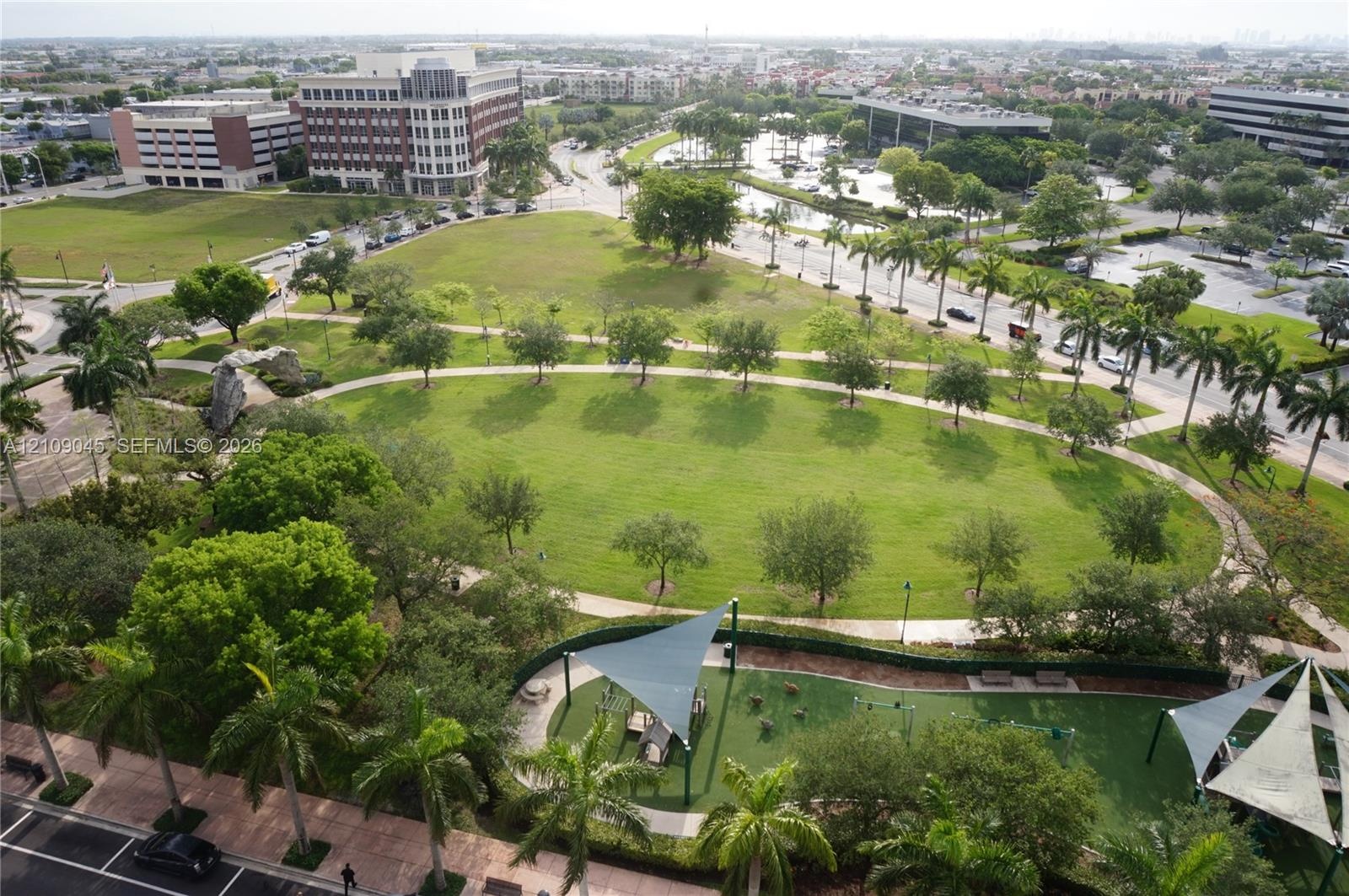 Expansive green space featuring mature trees, paved walking paths, curated landscaping, and shaded recreation structures