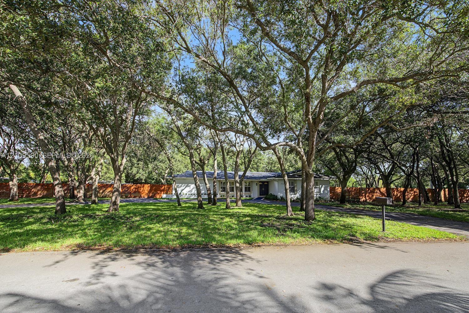 Single-story residence featuring a paved driveway, mature shade trees, expansive front lawn, and wood-paneled privacy fencing