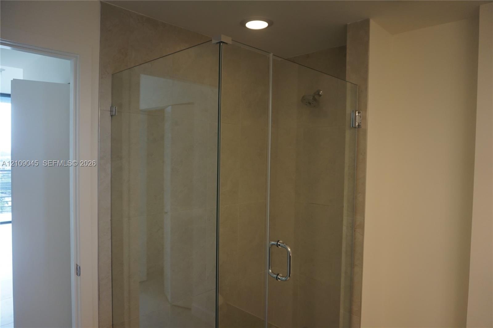 Shower enclosure with seamless glass panels, a polished metal handle, neutral-toned tiled surround, and a recessed ceiling light