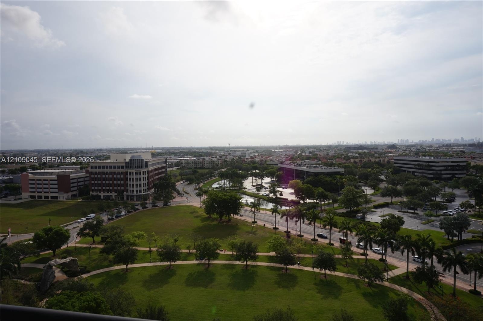 Elevated perspective featuring landscaped green space, mature palm trees, a central pond, and a horizon cityscape