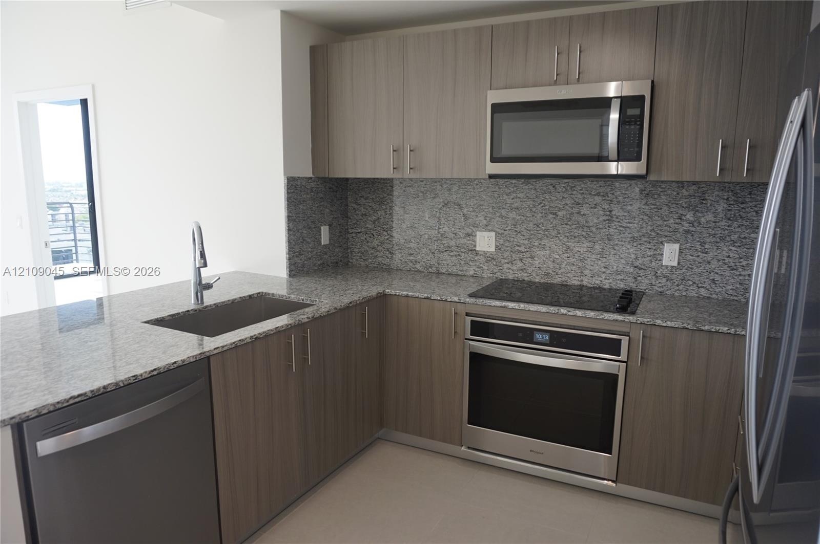 Kitchen featuring flat-panel wood-finish cabinetry, speckled stone countertops, a full-height stone backsplash, and stainless steel appliances includi