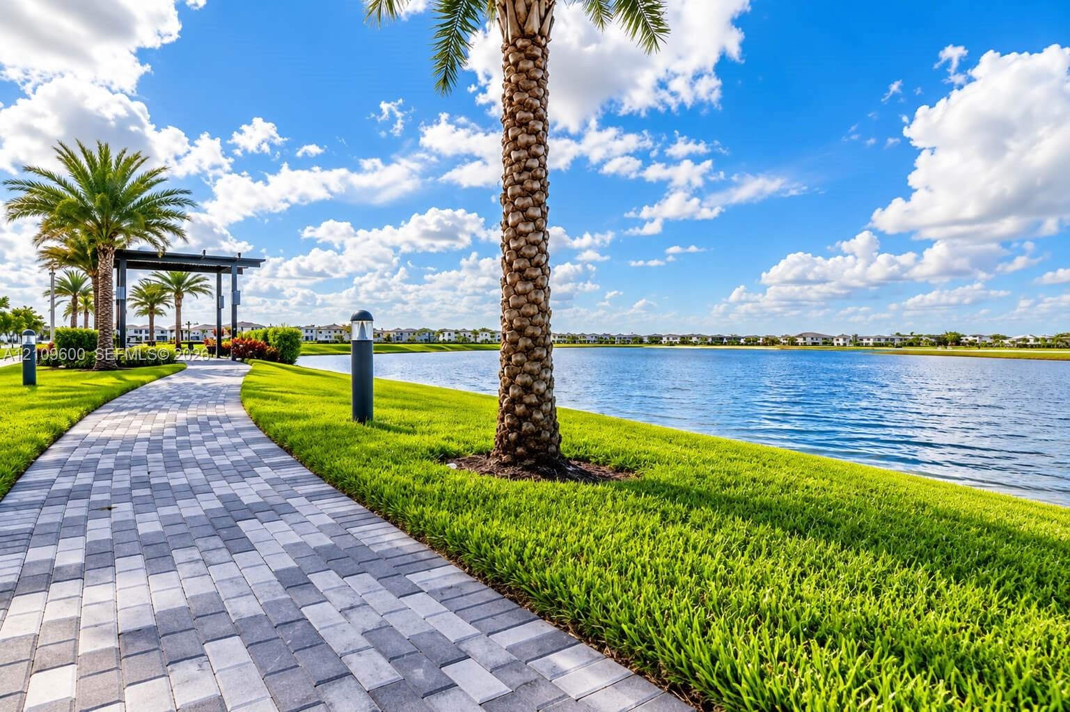 Paved stone walking path bordered by manicured lawn, lakefront frontage, mature palm trees, and an architectural metal pergola