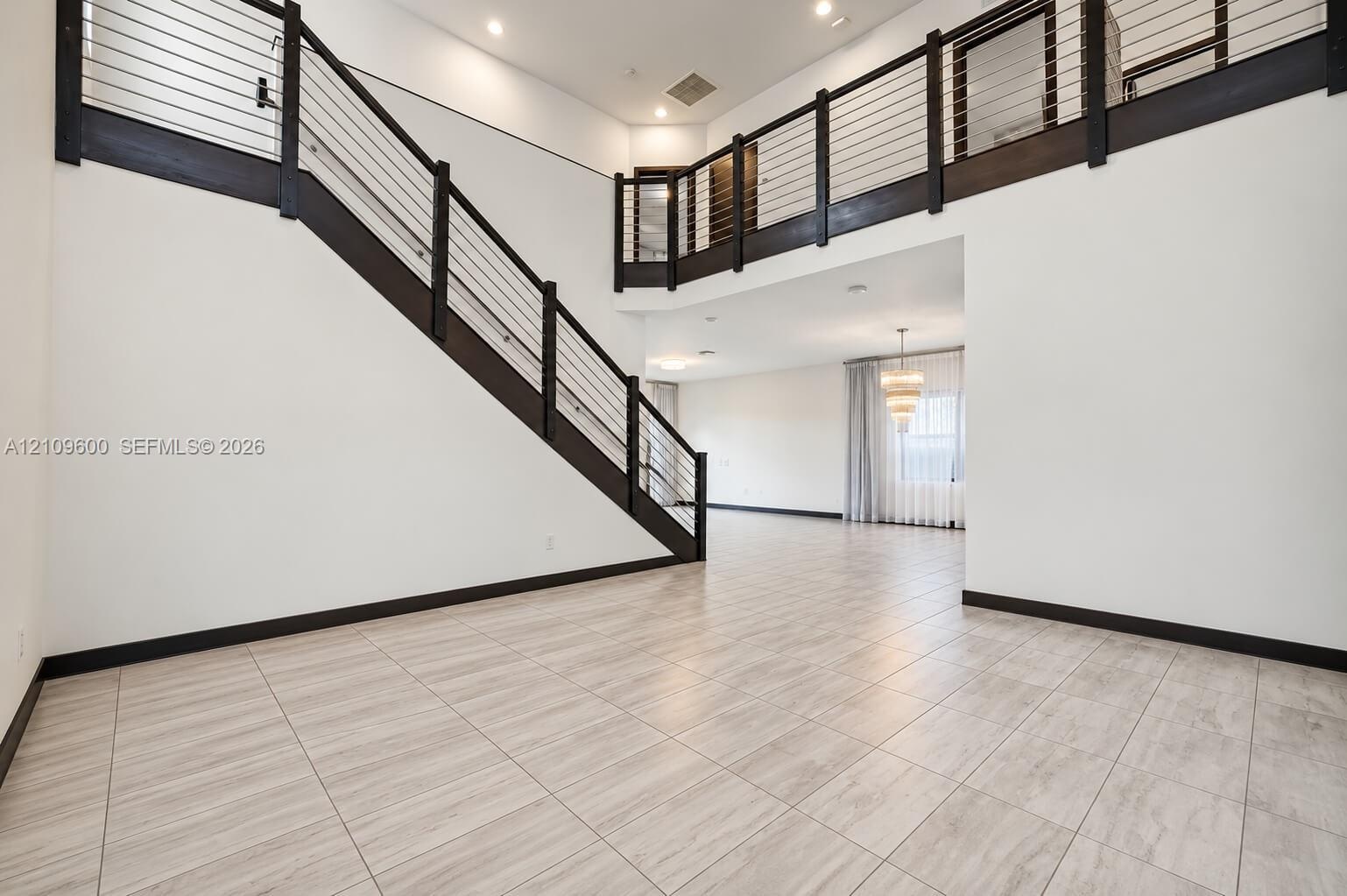Double-height ceiling, open-concept floor plan, staircase with metal horizontal railing, balcony landing, and neutral tile flooring
