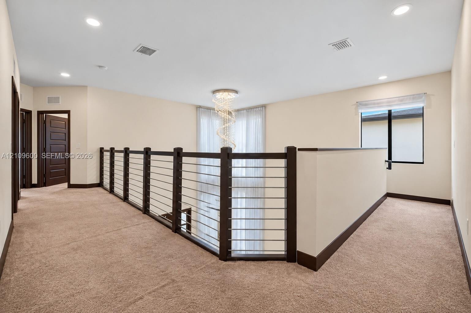Landing area featuring neutral carpeting, a dark-toned railing with horizontal metal balusters, a multi-tiered spiral chandelier, and solid-core panel