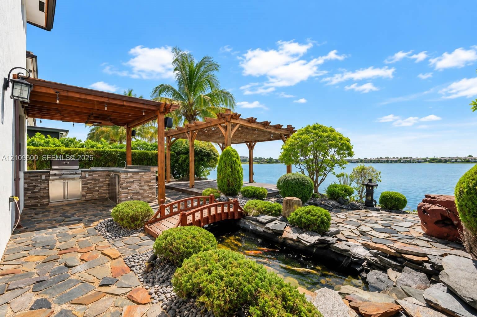 Waterfront outdoor living space featuring a stone patio, covered pergola, built-in stainless steel outdoor kitchen, and a decorative koi pond with a w