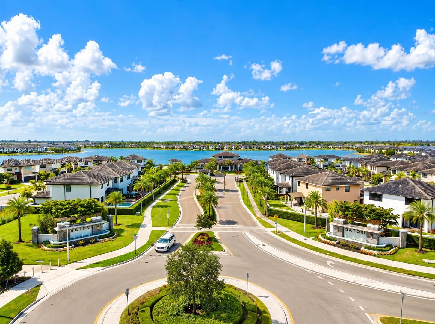 Aerial view of residential community featuring a paved roundabout, manicured landscaping, concrete sidewalks, multi-story residences, and a large lake