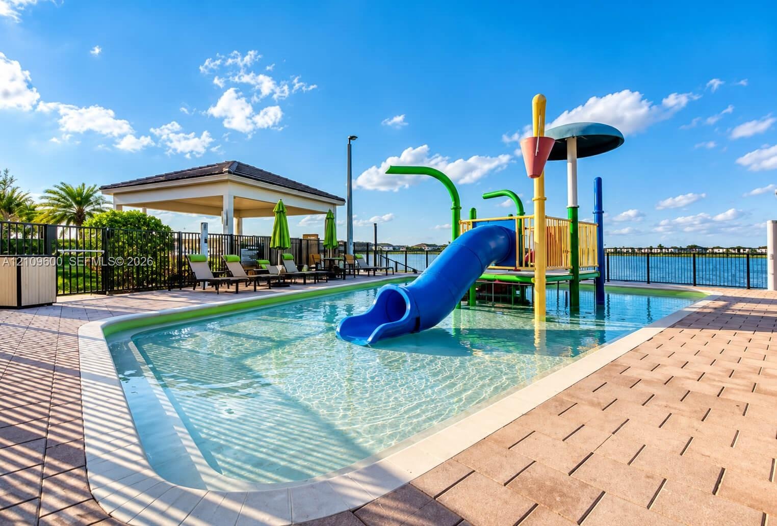 Outdoor pool with integrated water features and slide, paver-stone decking, and covered pavilion, situated against a waterfront backdrop