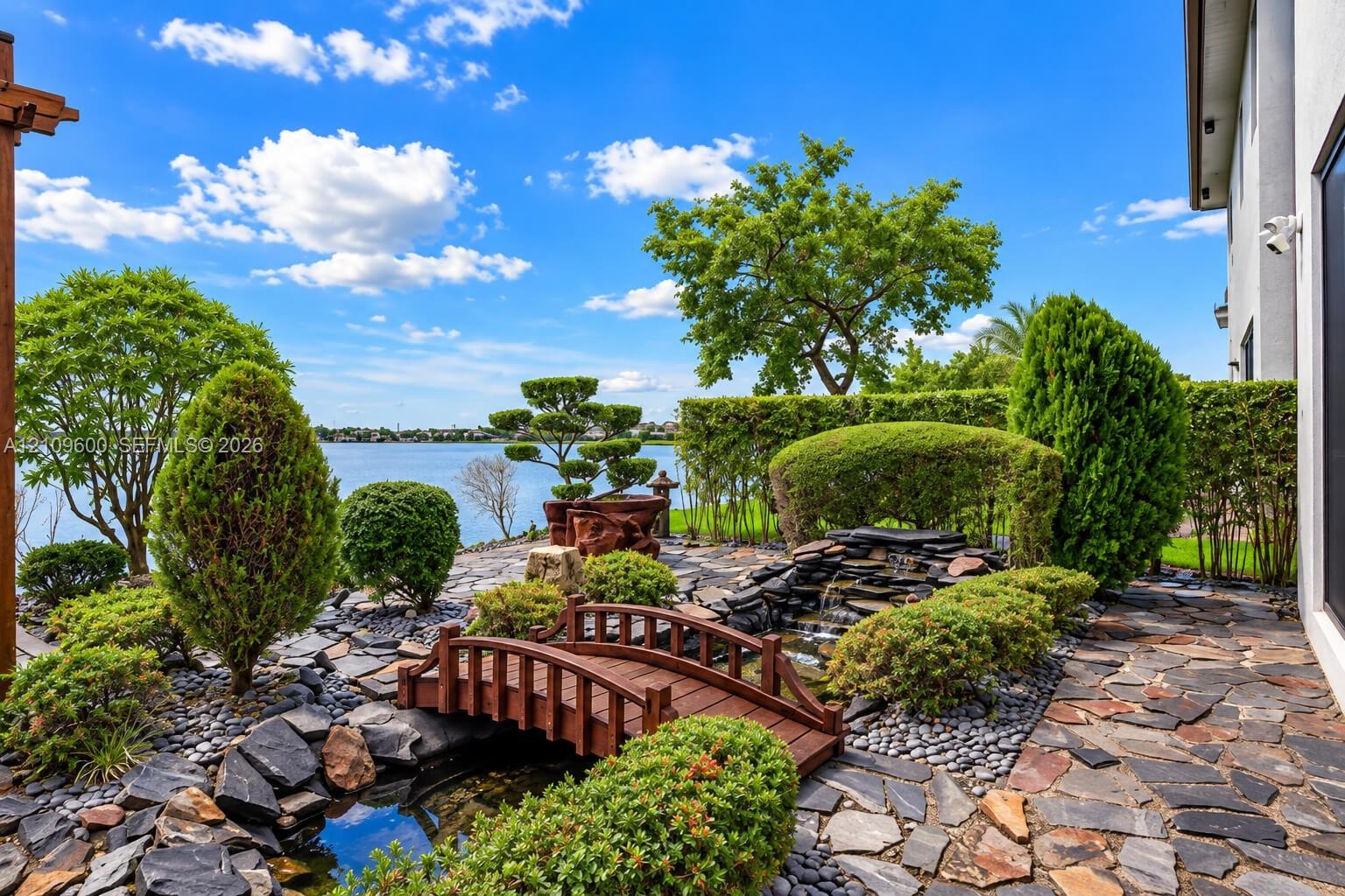 Landscaped backyard featuring a stone water feature, a wooden arched footbridge, flagstone hardscaping, and mature sculpted shrubbery