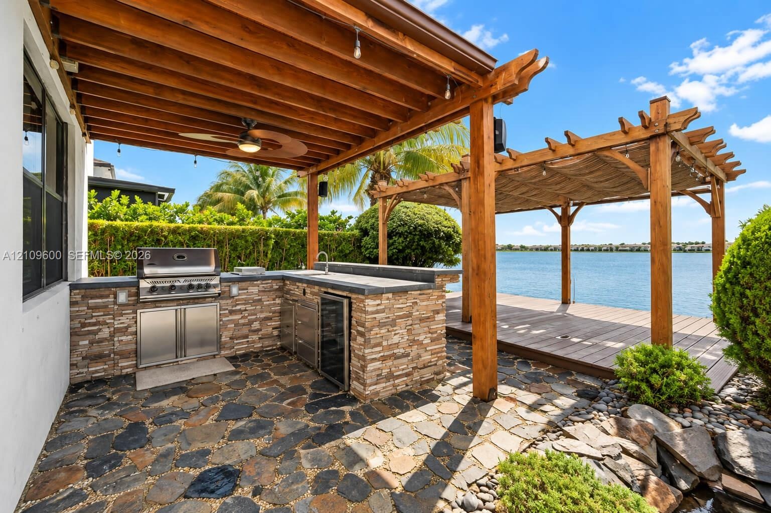 Outdoor kitchen featuring natural stone masonry, stainless steel built-in grill, and beverage refrigerator under a wood pergola, complemented by a sto