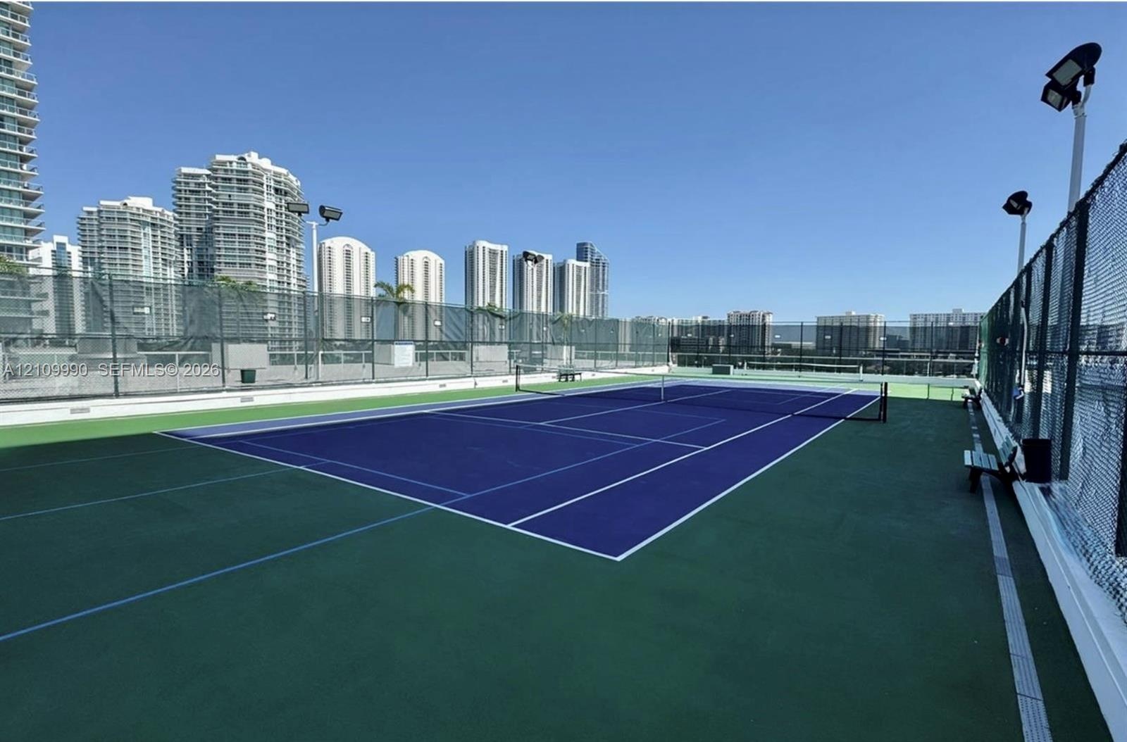 Rooftop tennis court featuring a blue and green multi-surface finish, perimeter chain-link fencing, and elevated stadium-style lighting