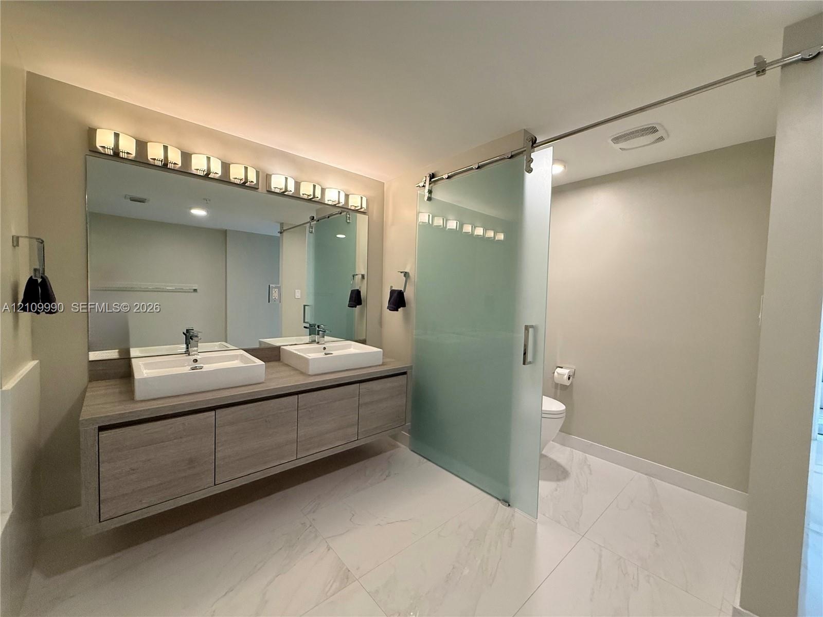 Floating wood-finish vanity with dual rectangular vessel sinks, expansive wall-to-wall mirror, oversized multi-bulb light fixture, and frosted glass s