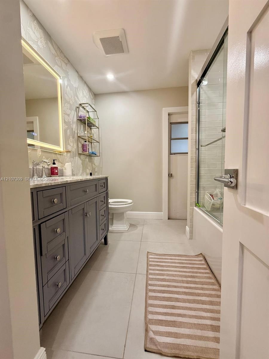 Bathroom featuring a grey shaker-style vanity with stone-surface countertop, LED-backlit mirror, large-format floor tile, and a shower-tub combination