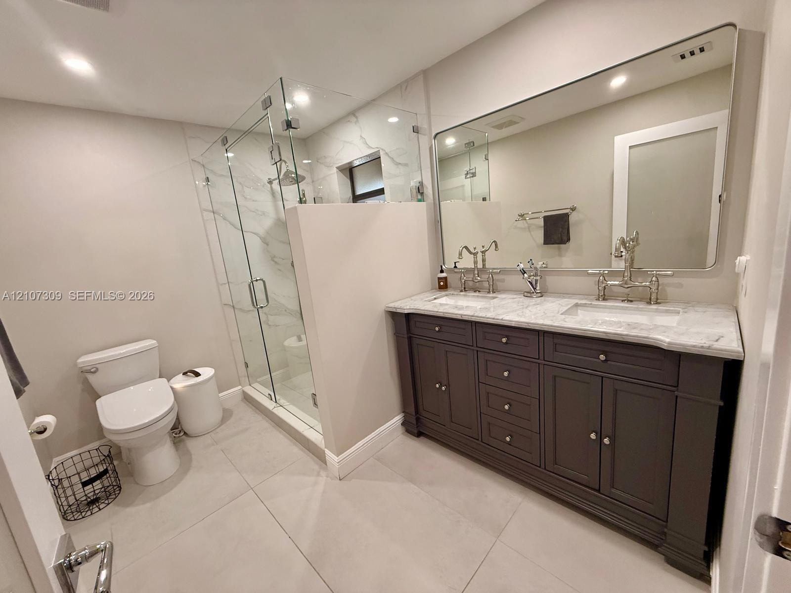 Dual-sink vanity with stone countertop and metallic fixtures, large wall-mounted mirror, walk-in glass-enclosed shower featuring stone-finish wall til