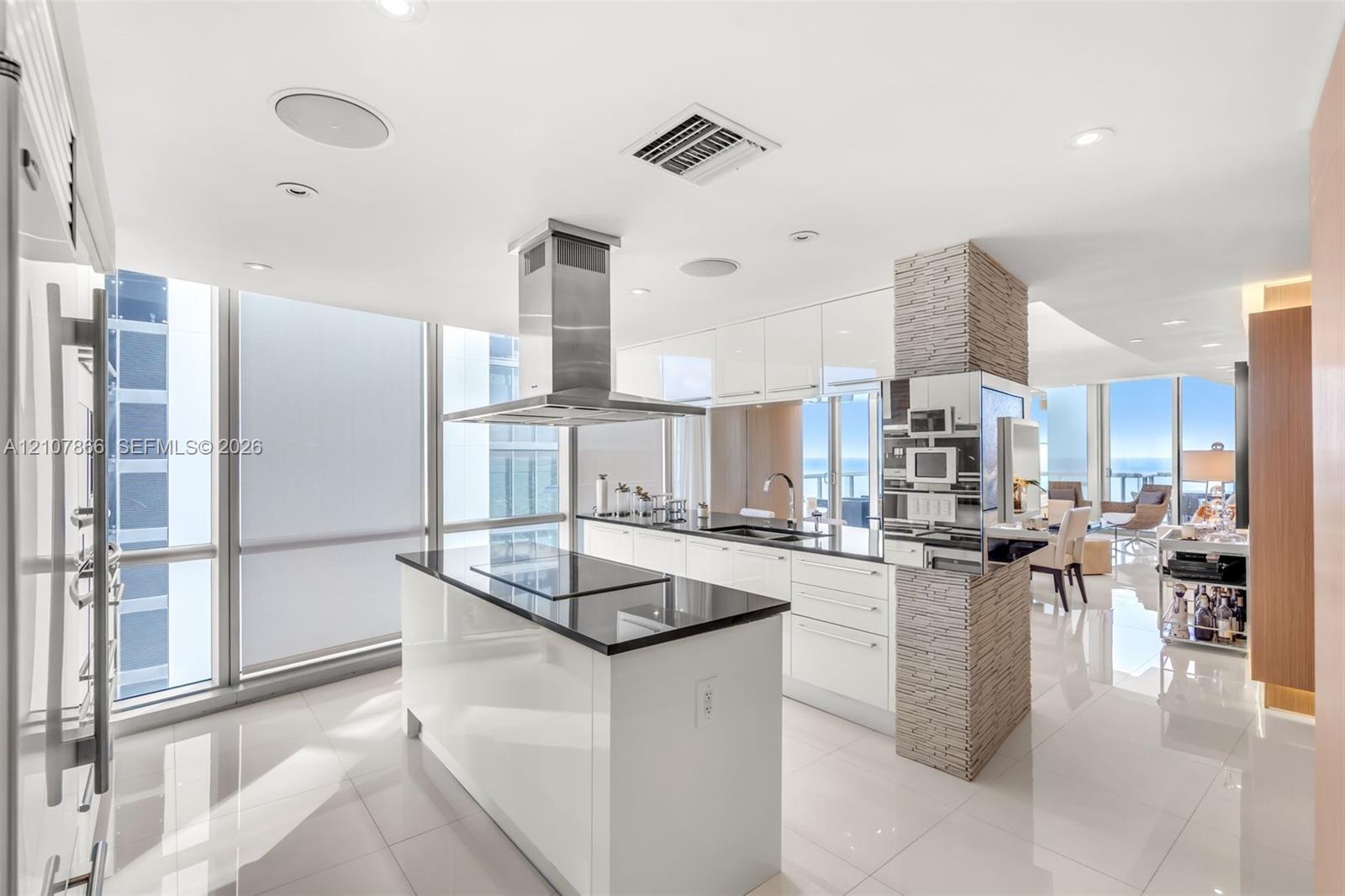 Contemporary kitchen featuring a center island with a sleek cooktop, stainless steel chimney-style range hood, high-gloss cabinetry, floor-to-ceiling 
