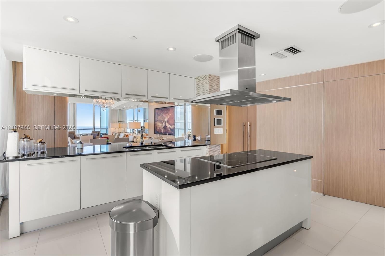 Modern kitchen featuring white flat-panel cabinetry, dark stone countertops, stainless steel island-mounted range hood, built-in cooktop, and wood-fin