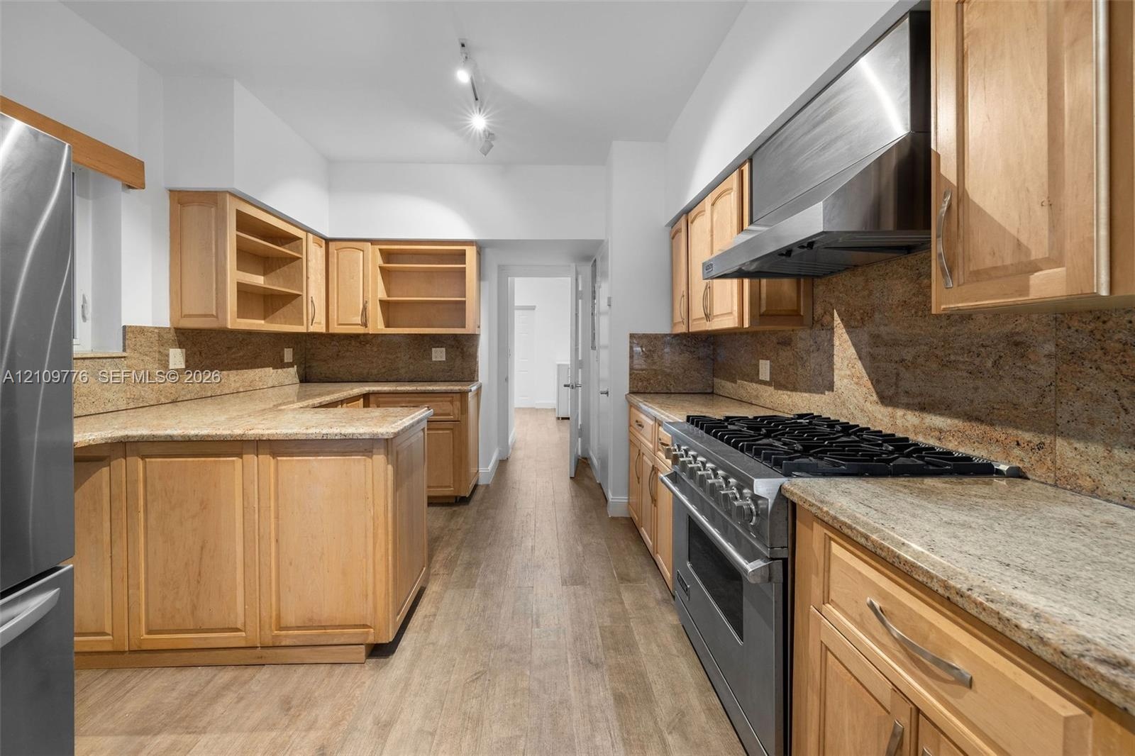 Kitchen featuring natural wood cabinetry, stone countertops, stone backsplash, stainless steel gas range with overhead vent hood, and wood-finish floo