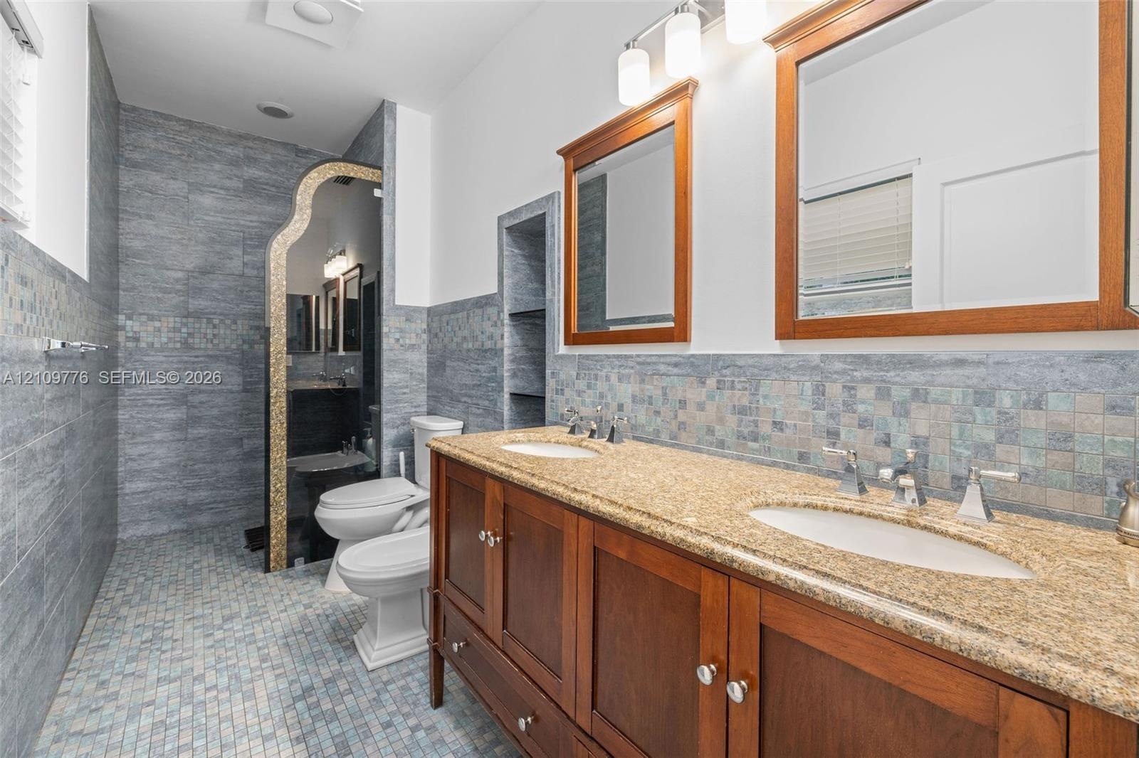 Dual-sink vanity with wood cabinetry, stone-finish countertop, decorative mosaic tile backsplash, built-in shelving, and framed wall mirrors