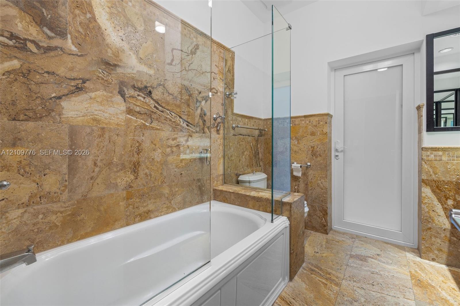 Full-height stone wall tiling, white paneled bathtub, frameless glass partition, matching stone flooring, and white door with frosted glass panel