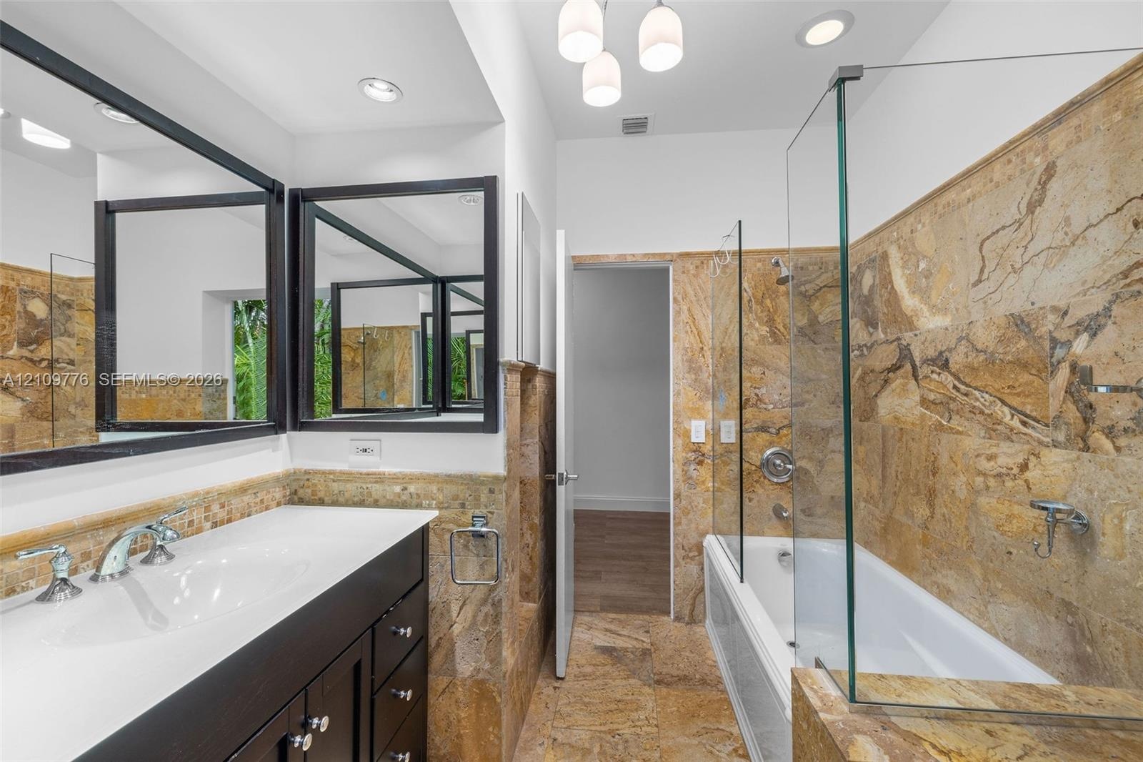 Bathroom featuring natural stone wall and floor tiling, dark-toned vanity with an integrated white countertop, frameless glass shower enclosure, and r