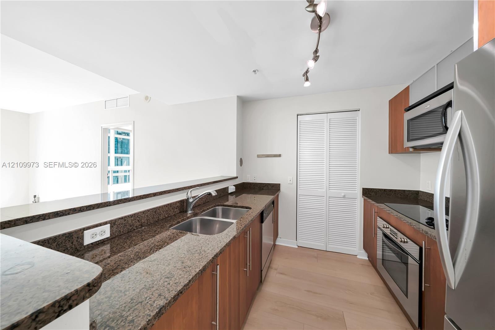 Kitchen featuring wood-finish cabinetry, stone countertops, stainless steel appliances, dual-basin sink, and track lighting