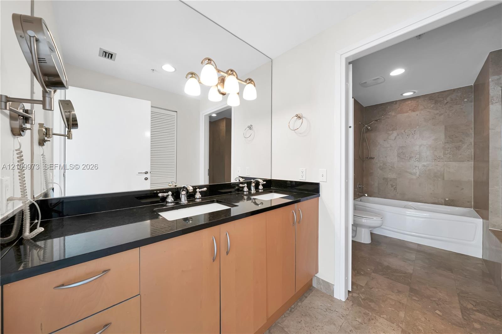 Dual-sink vanity with dark stone countertop, wood-finish cabinetry, full-width mirror, and multi-bulb vanity lighting, featuring a tile-surround batht