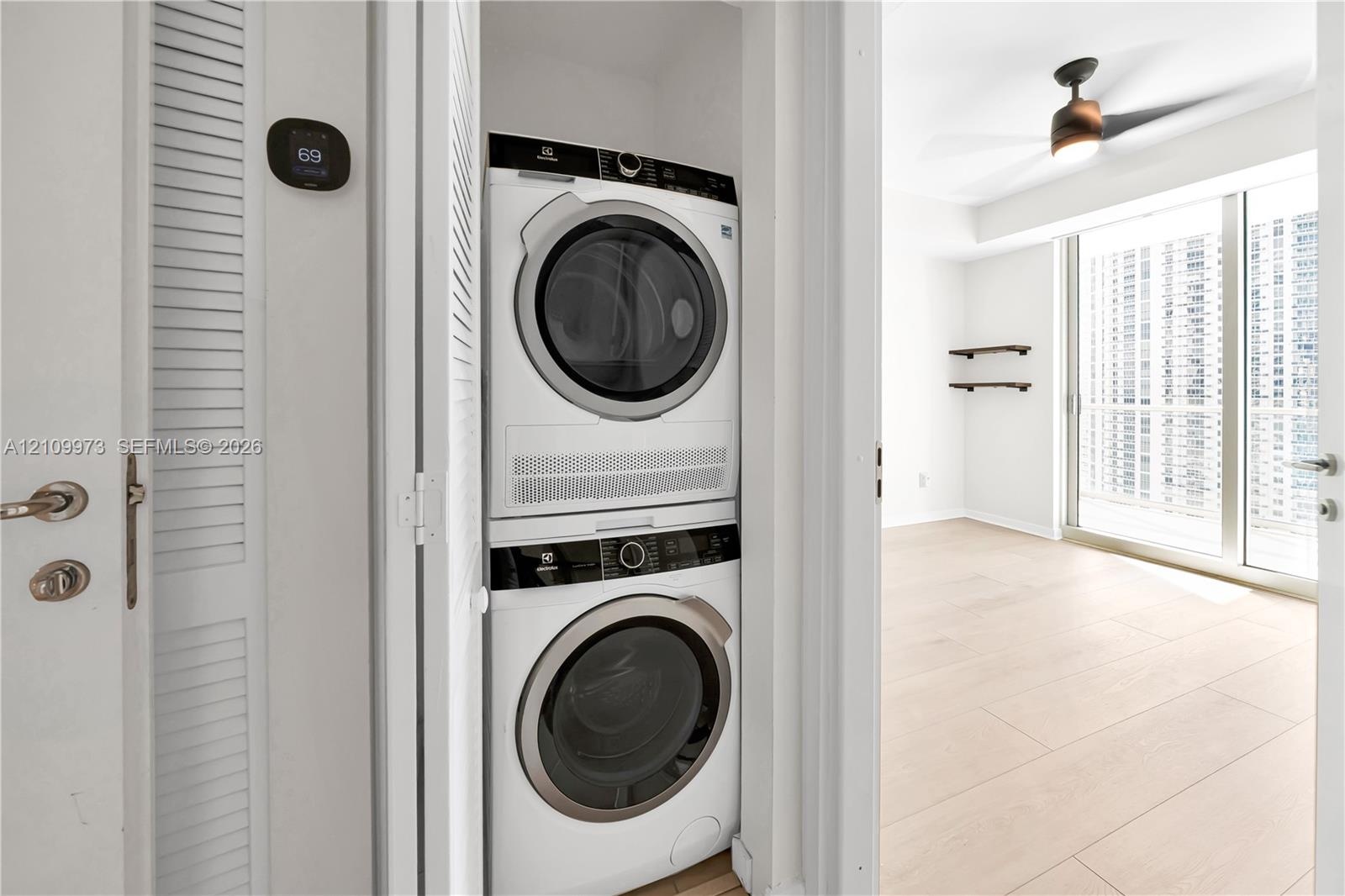 Stackable laundry unit featuring an Electrolux washer and dryer, digital wall-mounted thermostat, wood-finish flooring, ceiling fan with integrated li
