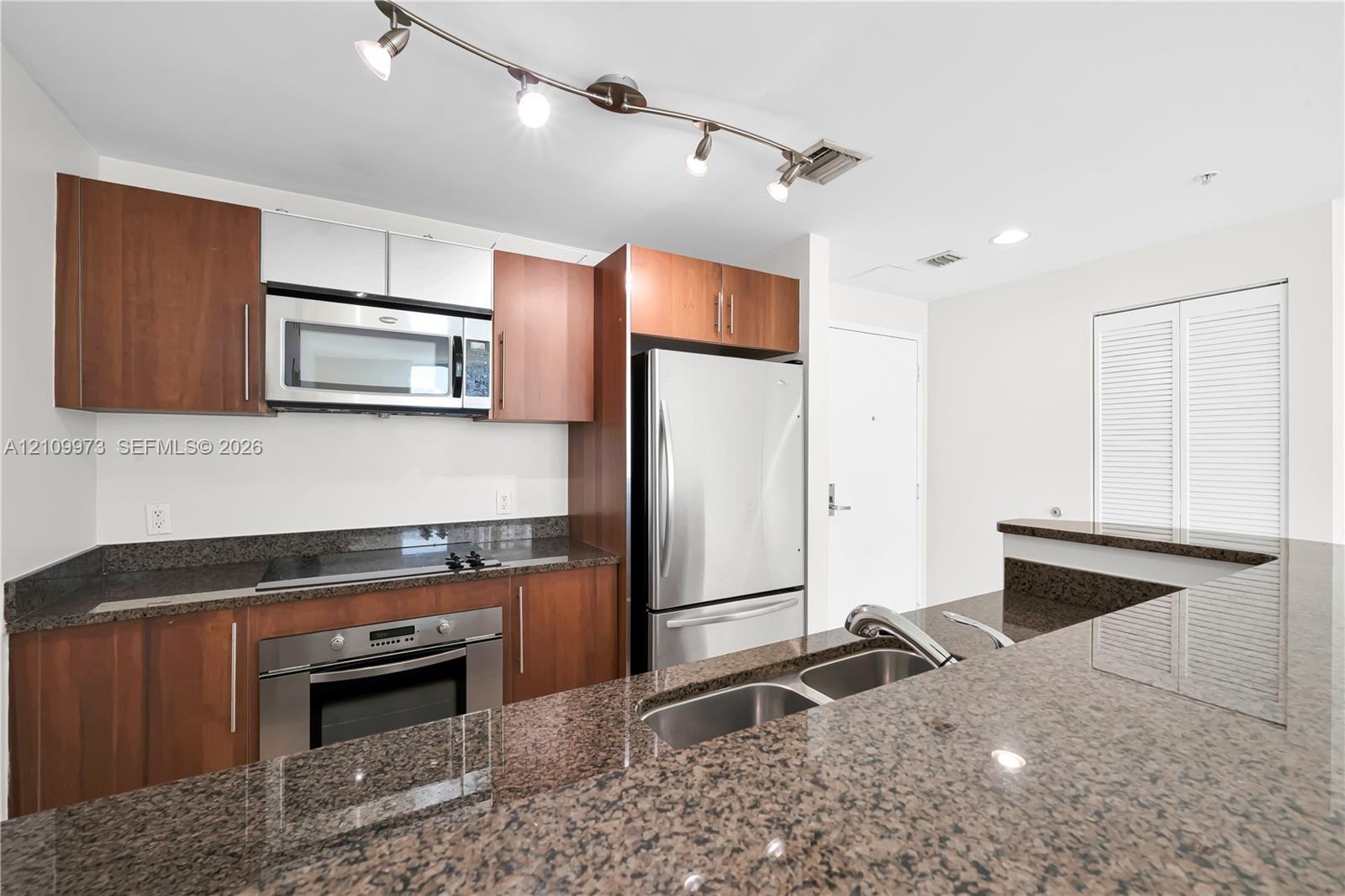 Kitchen featuring wood-finish cabinetry, stone countertops, stainless steel appliances, double-basin sink, and track lighting