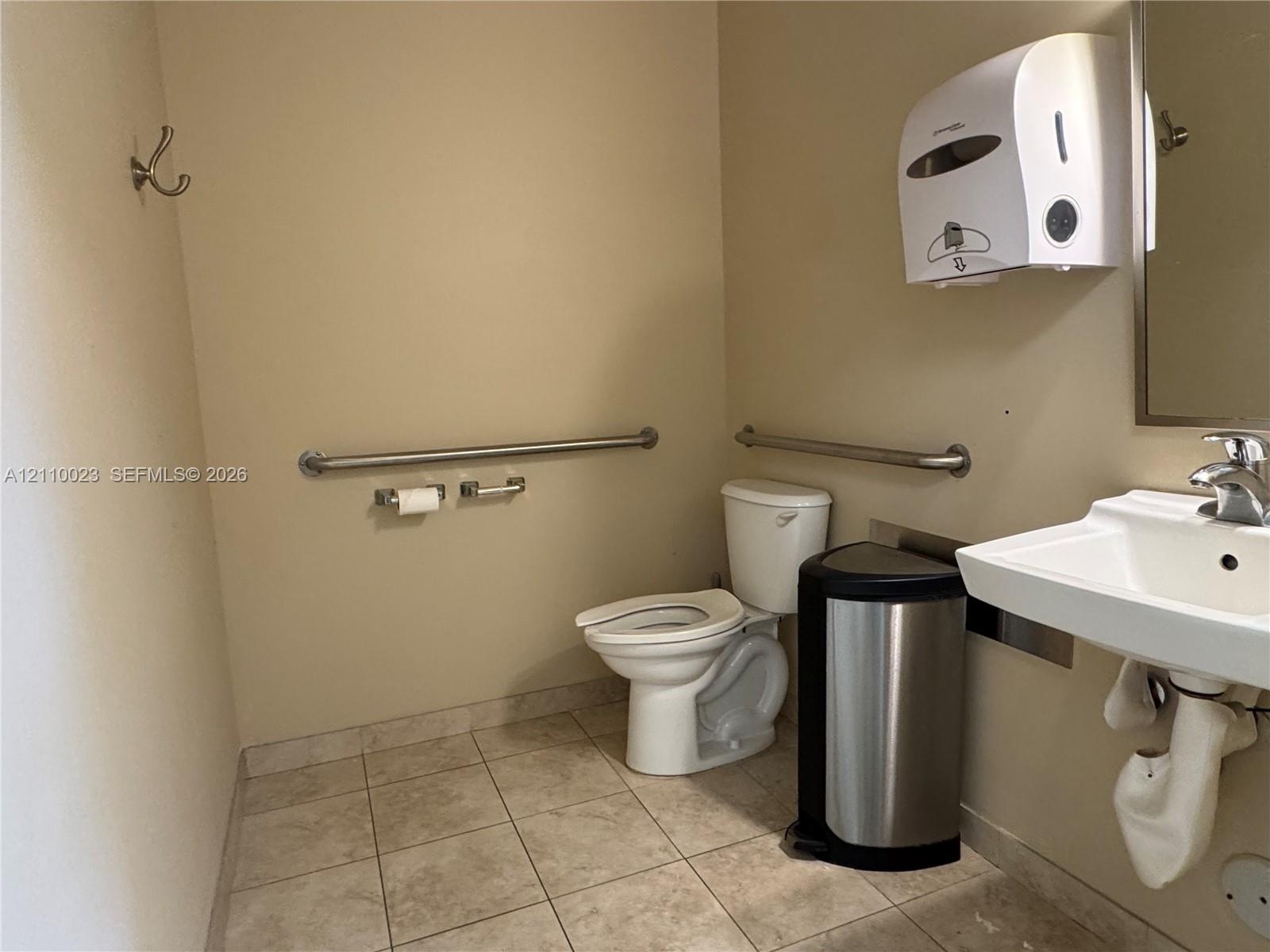 Restroom featuring tiled flooring, a wall-mounted sink with a chrome-finish faucet, an elongated porcelain toilet, and wall-mounted metal support bars