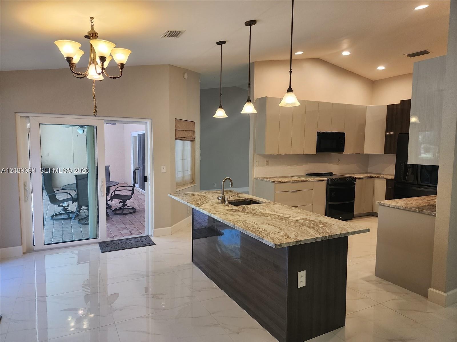 Kitchen featuring stone-topped island with undermount sink, flat-panel cabinetry, porcelain tile flooring, trio of pendant lights, and sliding glass d