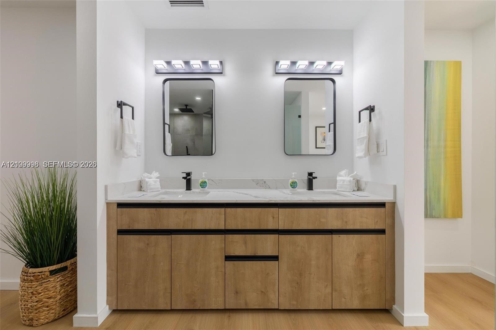 Double vanity featuring a stone-surface countertop, dual rectangular sinks with matte black faucets, light wood-finish cabinetry with linear pulls, an