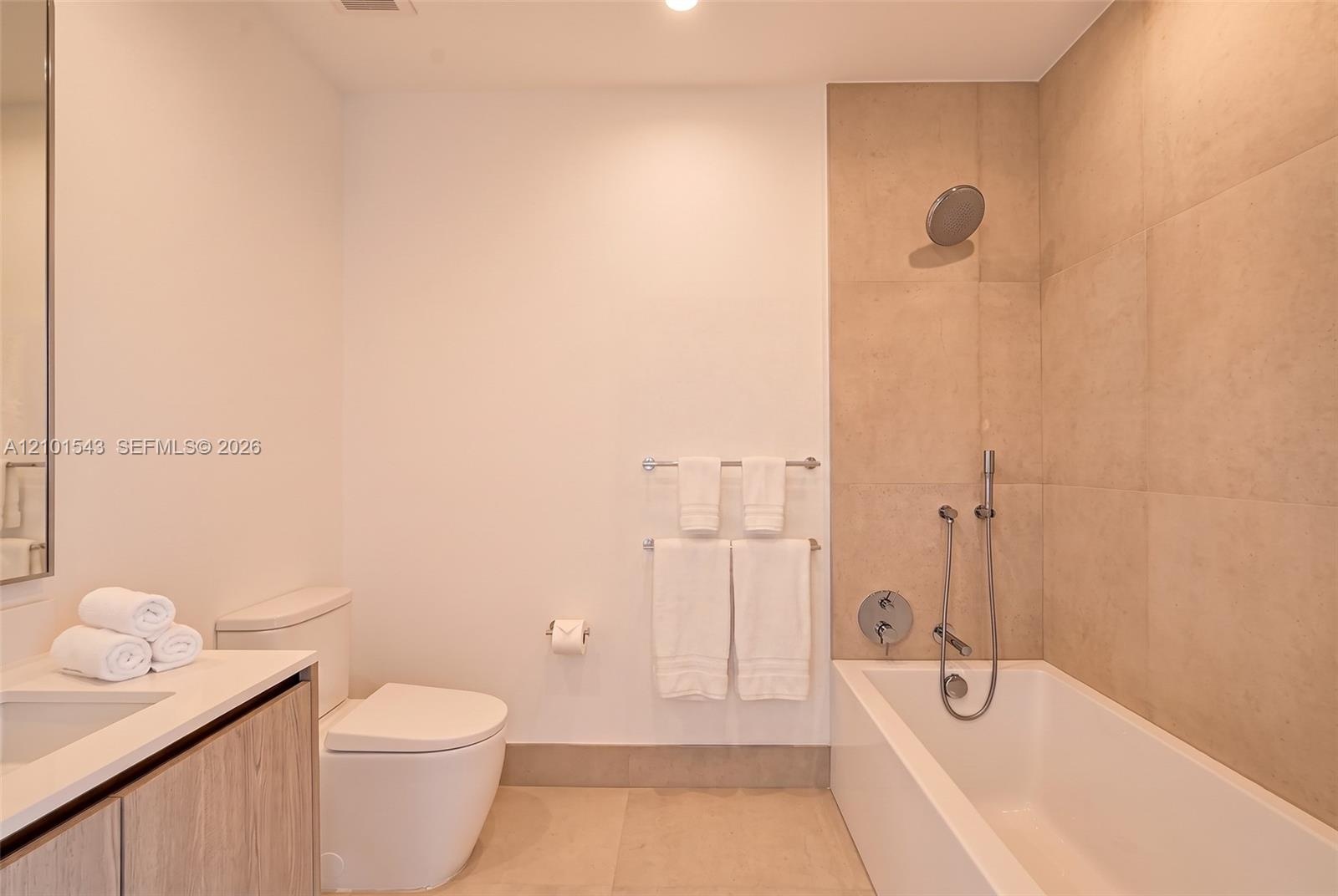 Bathroom featuring a wood-finish vanity, large-format neutral wall tile, integrated tub and shower combination with handheld fixture, and recessed lig