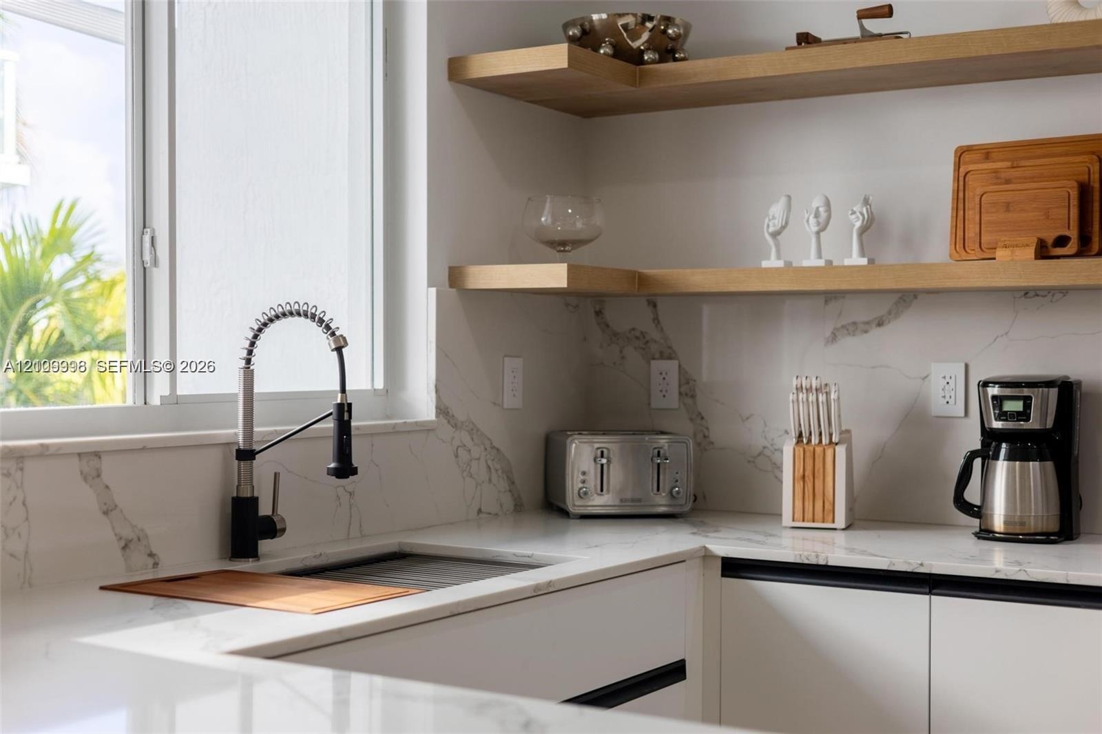 White cabinetry with black recessed pulls, stone-composite countertops with matching full-height backsplash, wall-mounted wood floating shelving, and 