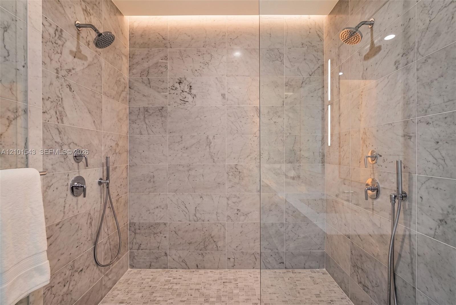 Walk-in shower featuring large-format stone wall tiling, mosaic stone flooring, dual shower heads with handheld sprayers, a frameless glass enclosure,