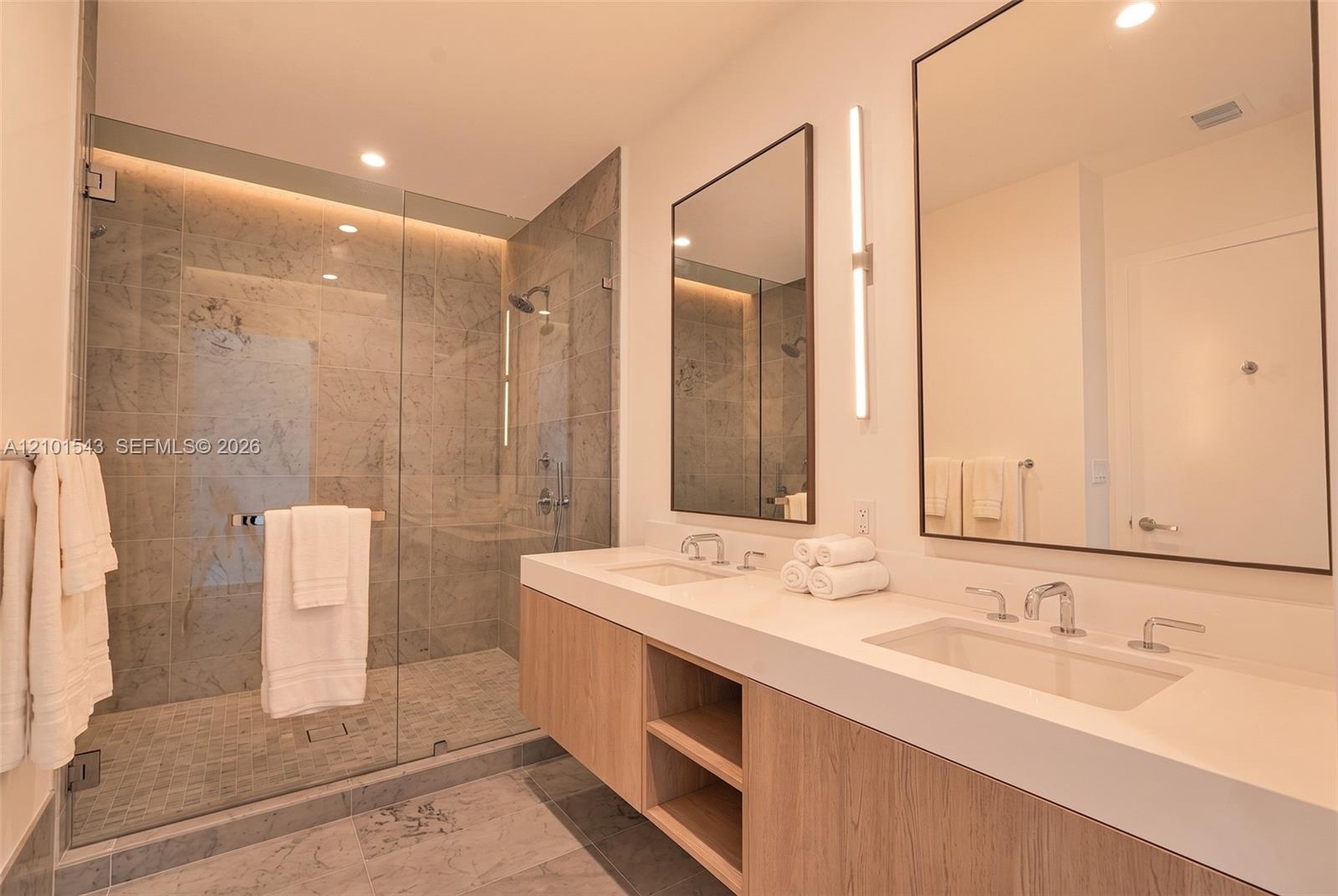 Floating dual-sink vanity with a solid surface countertop, glass-enclosed walk-in shower with stone tile surround, stone tile flooring, and integrated