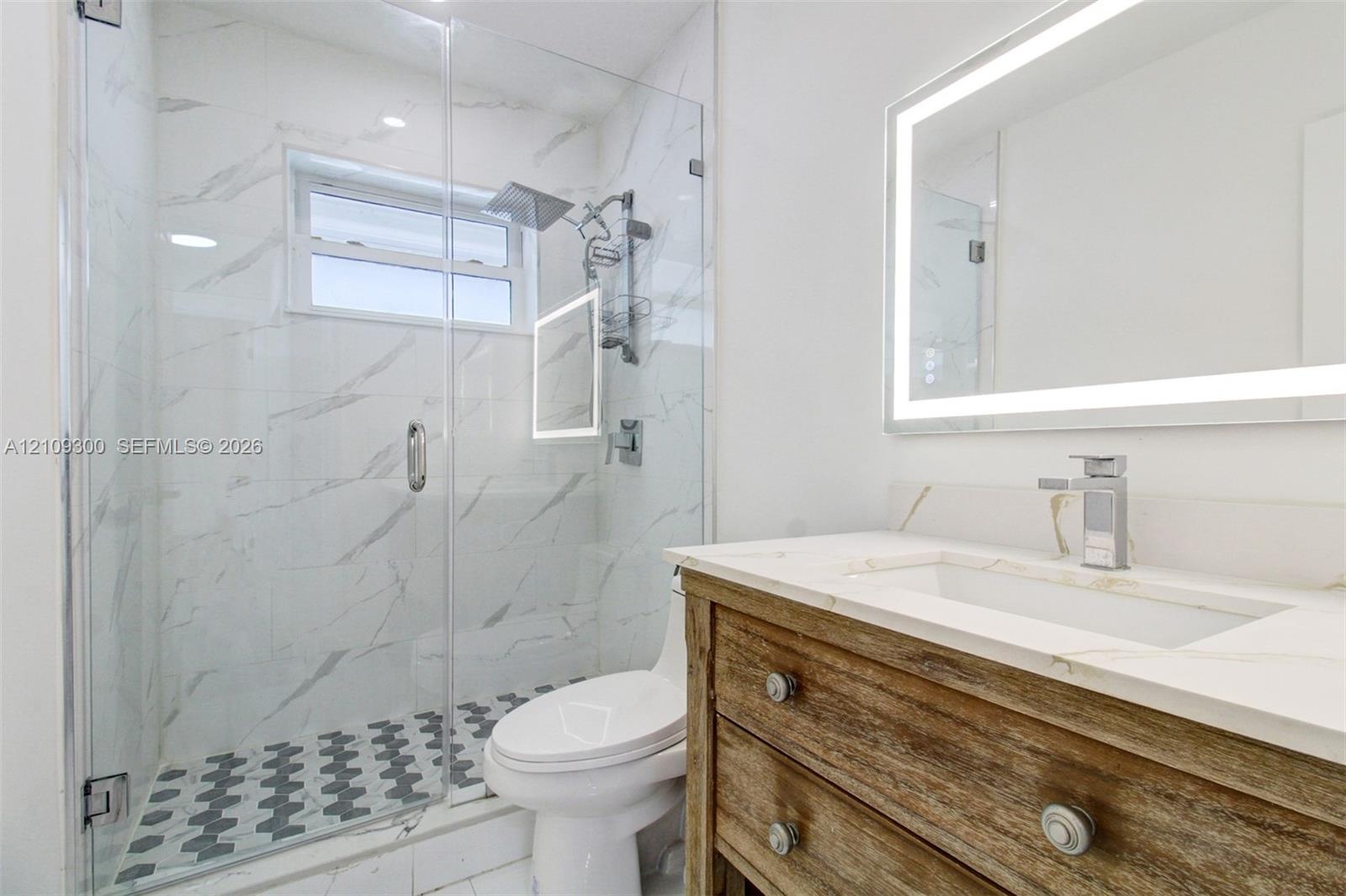 Bathroom featuring a wood-finish vanity with a stone countertop, an integrated rectangular sink, a frameless glass-enclosed shower with large-format t