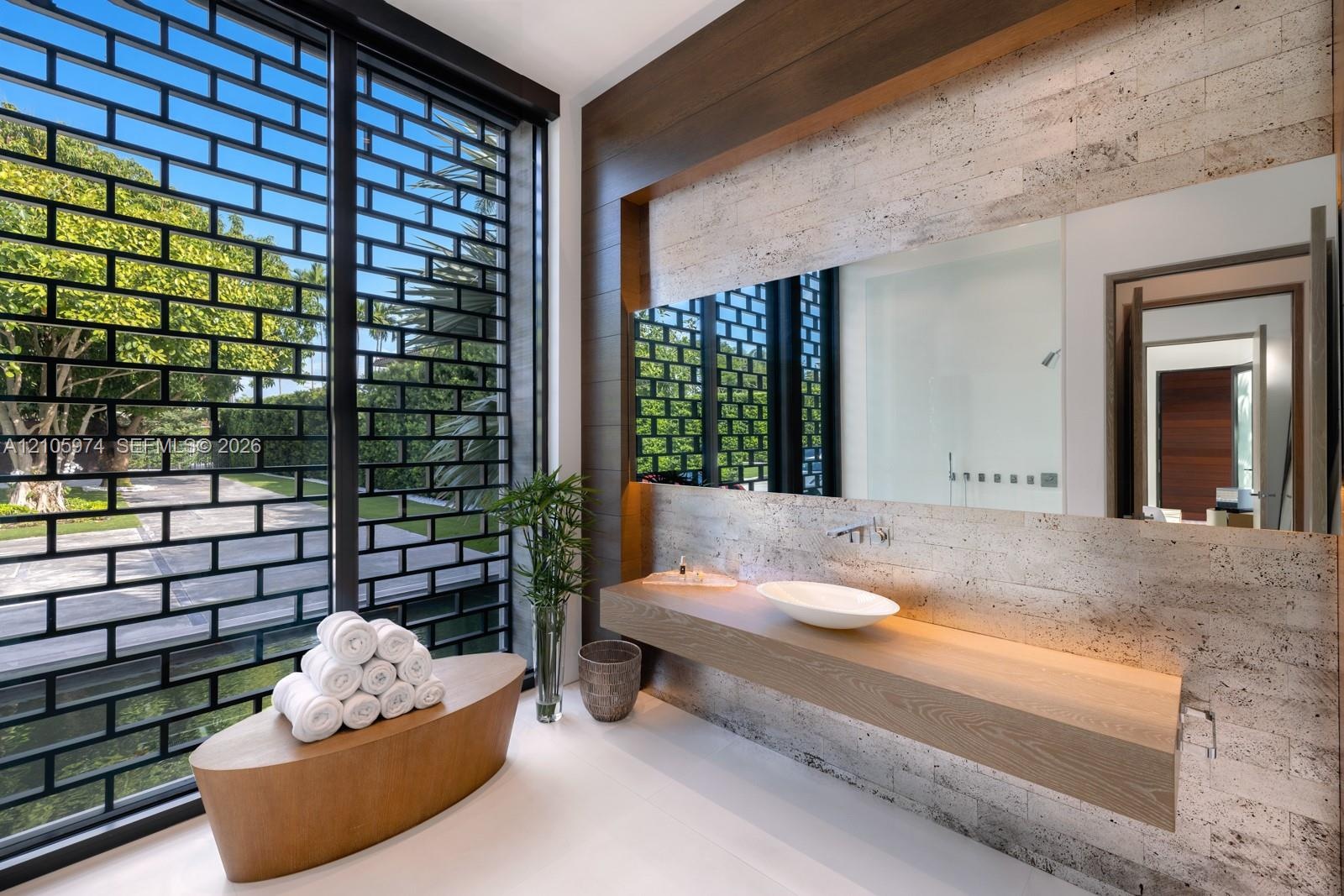 Floating wood-finish vanity with a white vessel sink, wall-mounted faucet, oversized frameless mirror, textured stone-tiled wall, and full-height exte