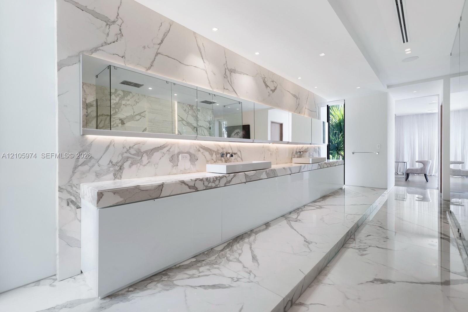 Large-format stone wall and floor tiling featuring prominent veining, integrated full-width vanity mirror with recessed lighting, and wall-mounted fau