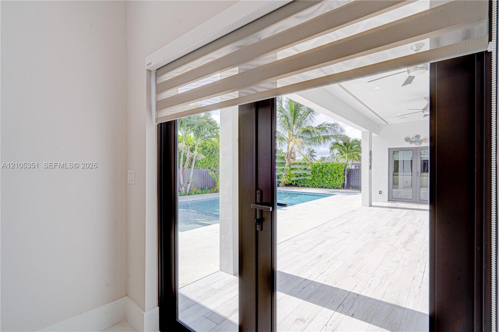 Dark-framed glass double doors featuring dual-layer roller shades, opening onto a covered patio with light-toned tile flooring, recessed ceiling light