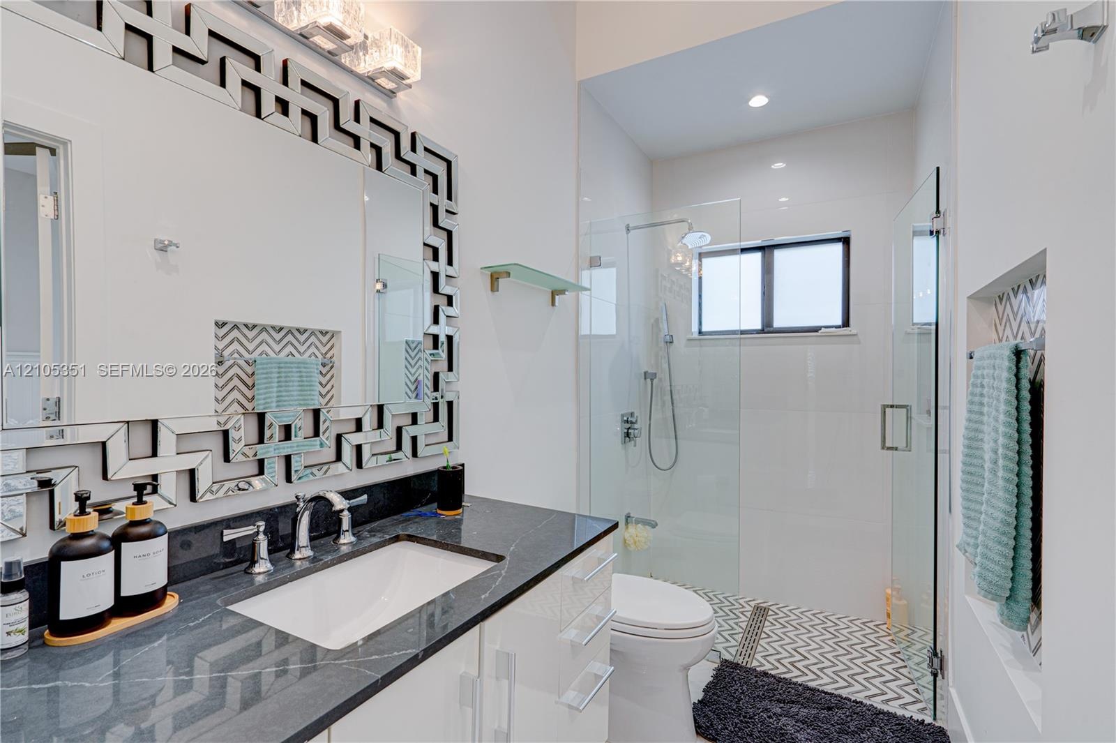 Glossy white cabinetry, dark stone countertop with an undermount sink, geometric mirror frame, walk-in glass-enclosed shower with chevron-patterned ti