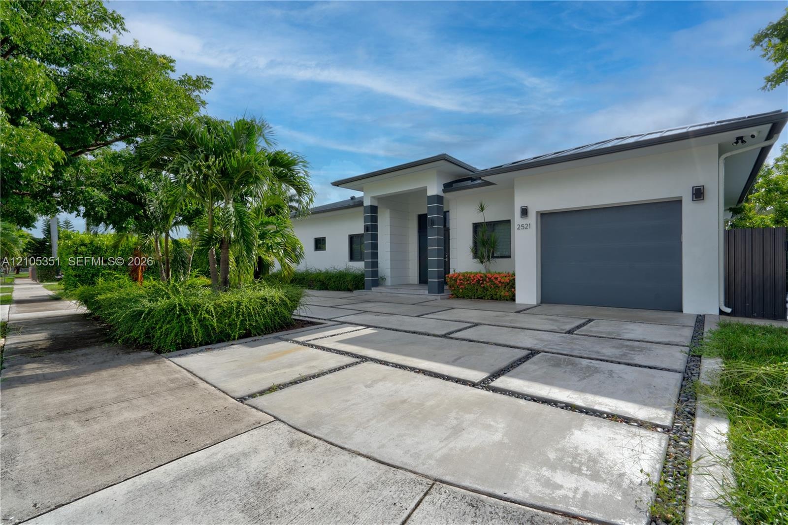 Modern exterior featuring a geometric concrete paver driveway, covered entryway with column detailing, attached garage, and landscaping with mature pa