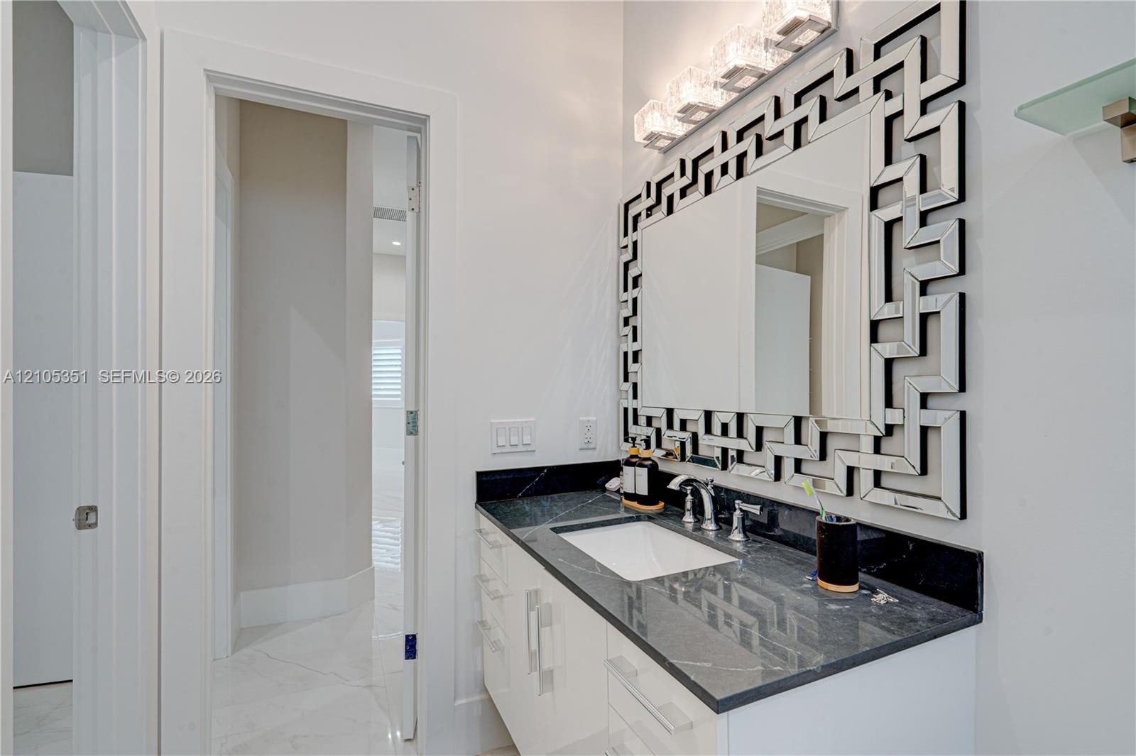 White vanity with dark stone countertop, undermount rectangular sink, chrome widespread faucet, geometric-patterned mirror, and four-light glass wall 