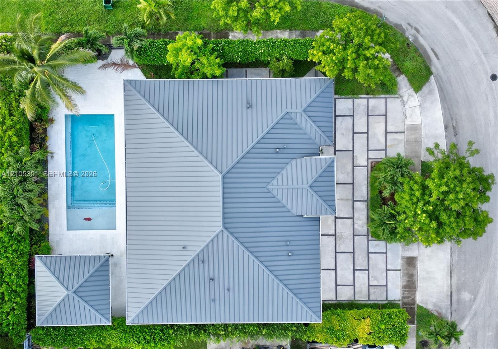 Aerial perspective featuring a metal roof, rectangular swimming pool, separate cabana structure, and expansive stone paver hardscaping bordered by mat