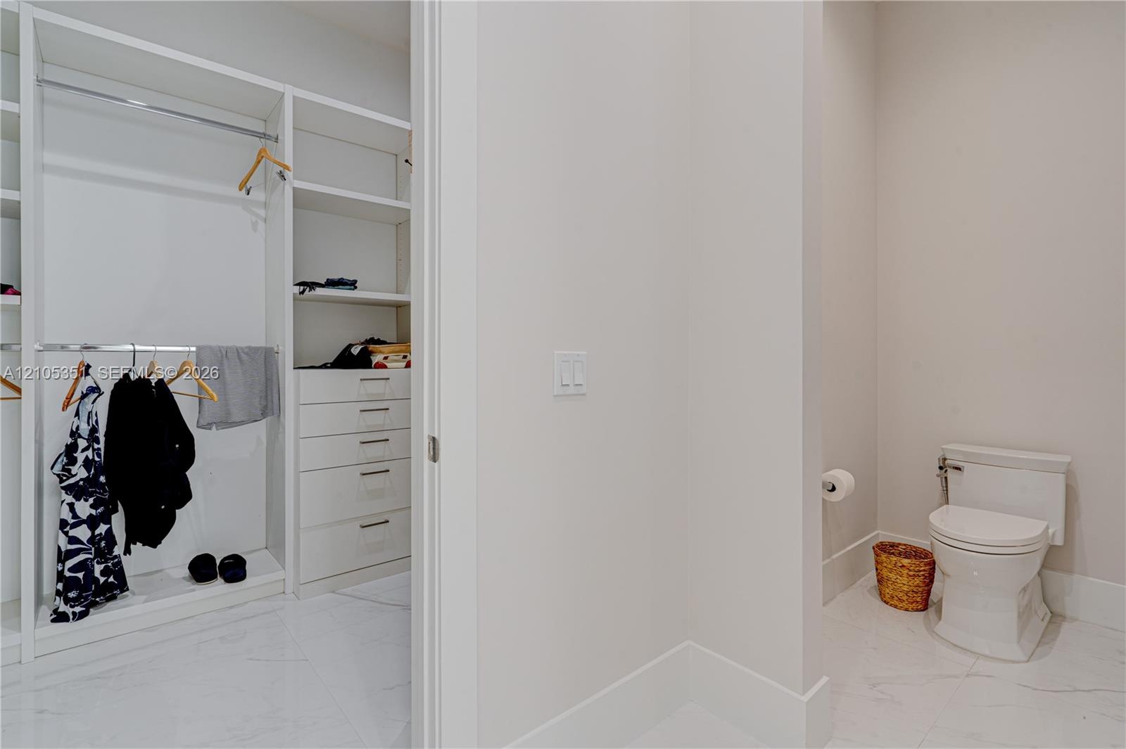 Custom walk-in closet featuring white built-in shelving and integrated drawer storage, paired with an enclosed water closet finished with stone-tile f