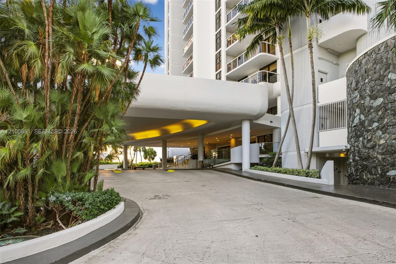 Covered porte-cochère entry featuring stone masonry accents, a concrete drive, and mature palm landscaping