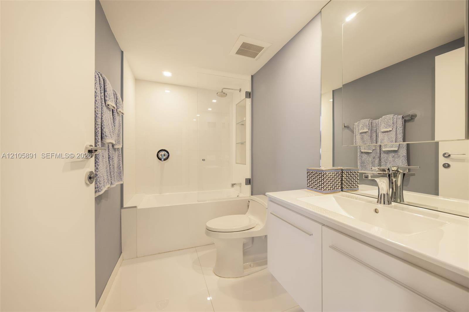 Bathroom featuring a white vanity with an integrated sink, a large wall-mounted mirror, a tub-shower combination with a glass panel, and polished tile