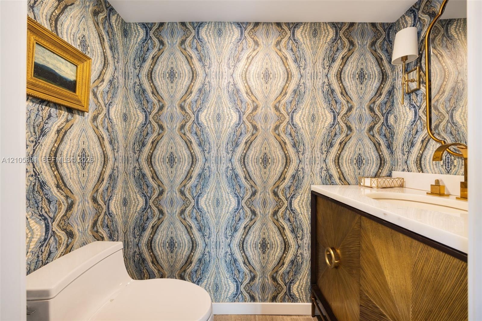 Powder room featuring patterned wallpaper, a wood-finish vanity with sunburst detailing, a stone-composite countertop, an integrated sink, and brass-f