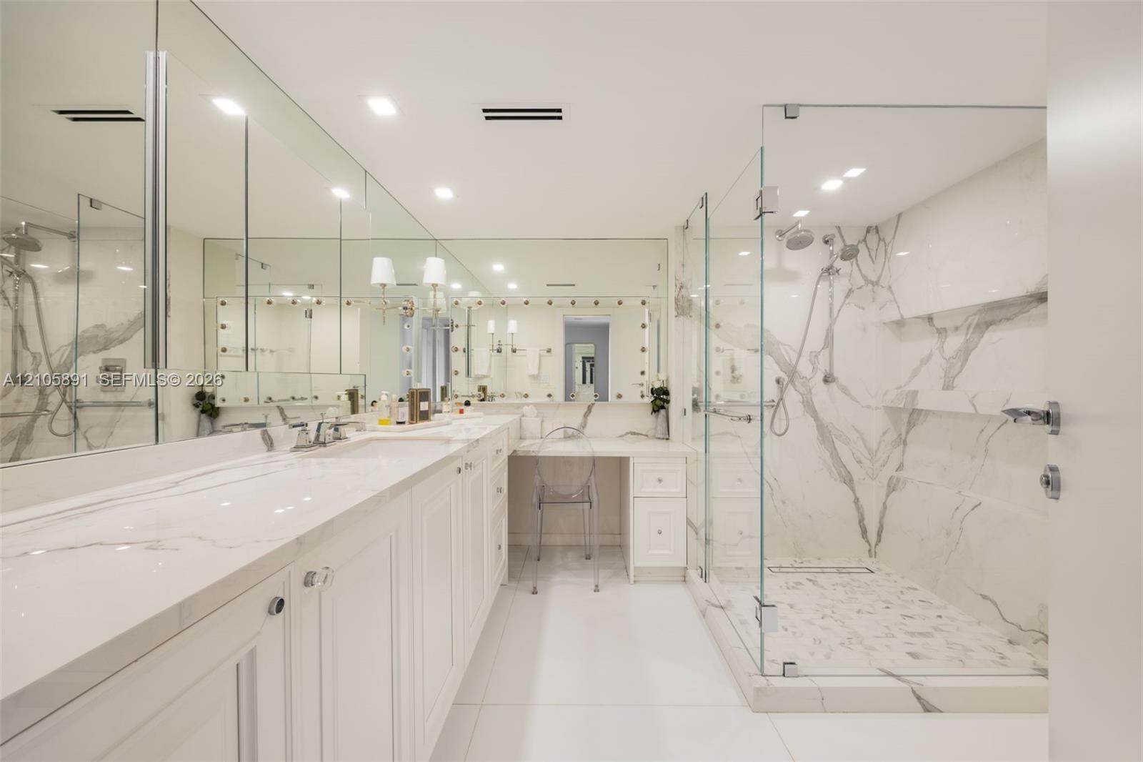 Dual-sink vanity with quartz countertop, expansive wall-to-wall mirrors, integrated makeup vanity station, and glass-enclosed walk-in shower featuring