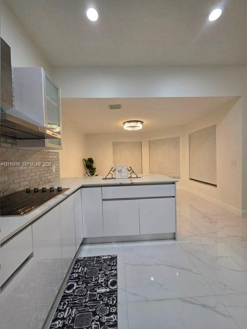 Kitchen featuring glossy white flat-panel cabinetry, light-toned stone-finish flooring, a textured stone-tile backsplash, and recessed overhead lighti