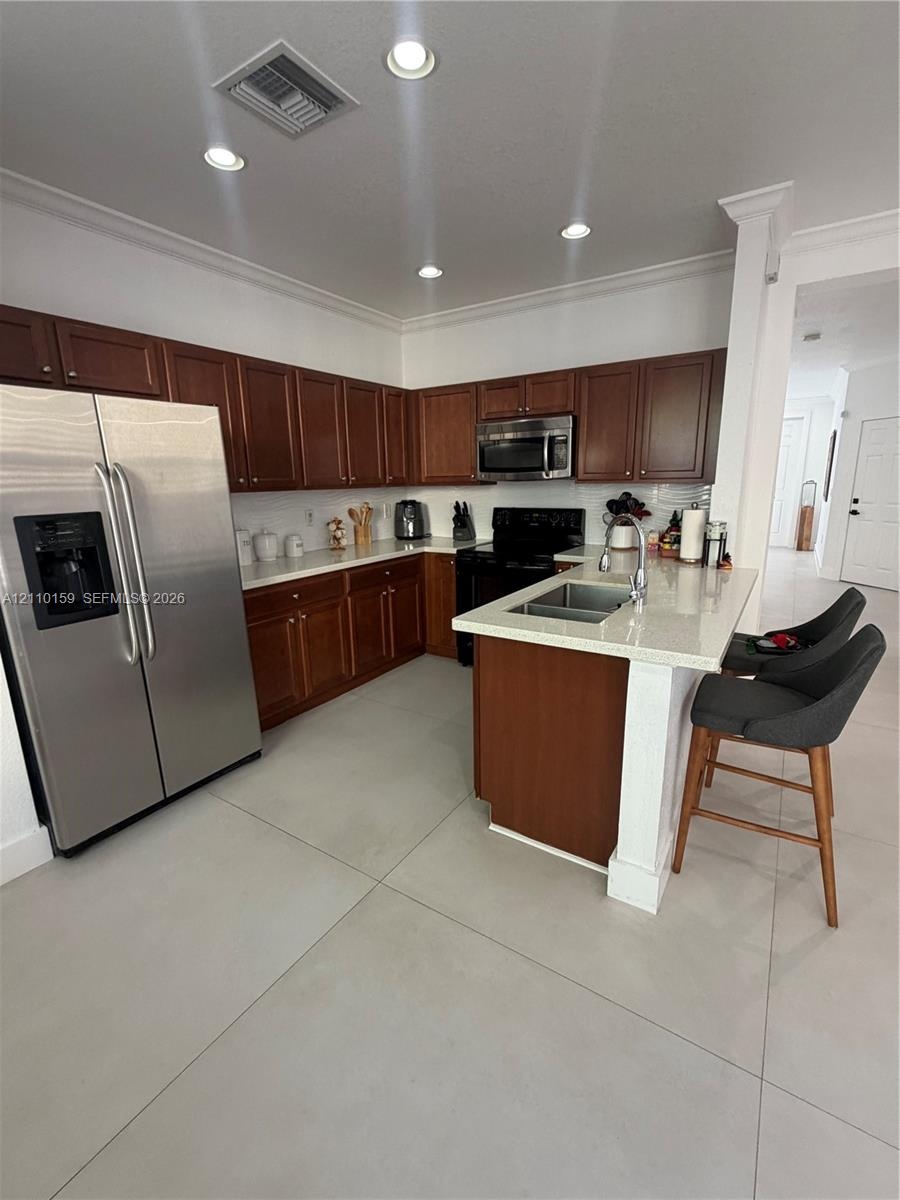 Kitchen featuring wood cabinetry, light-toned stone countertops, a center island with a dual-basin stainless steel sink, large-format tile flooring, a